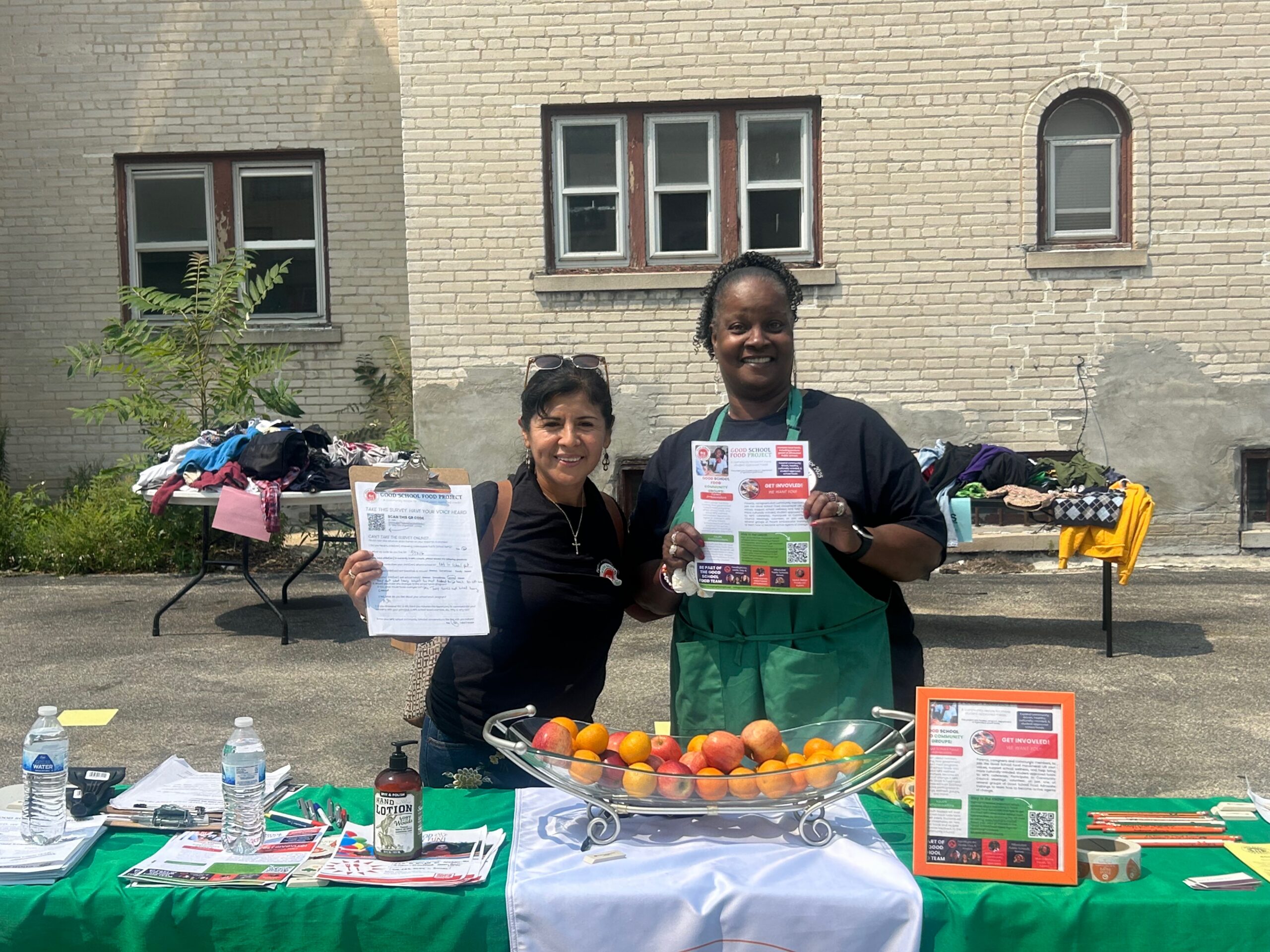 Two women at an outdoor event in front of a bowl of fruit, holding up a clipboard and a flyer.